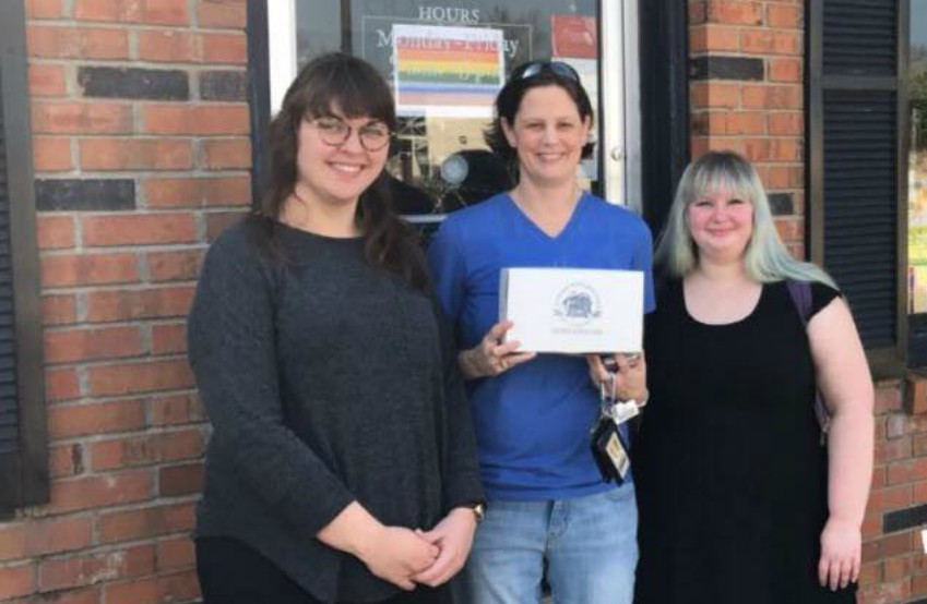 Three women stand in front of bakery, the middle one is holding up a box of cupcakes there is a rainbow flag in the window behind them